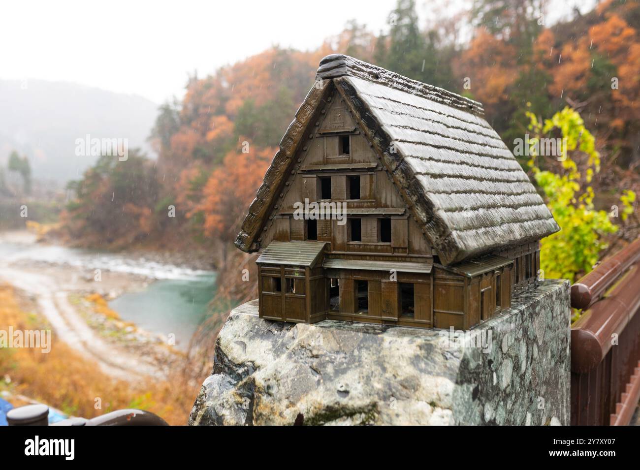 Una dettagliata miniatura di una tradizionale casa in legno si trova su una sporgenza di pietra, con un fiume e vivaci alberi autunnali sullo sfondo. Foto Stock