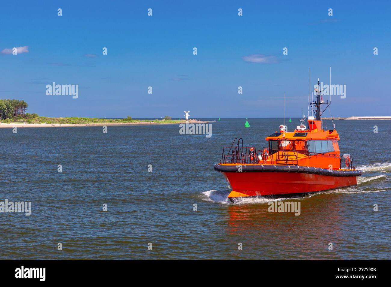 Un pilota di barca arancione che naviga alla foce del fiume Swina a Swinoujscie, in Polonia, con il faro Stawa Mlyny visibile sulla frangiflutti sullo sfondo Foto Stock