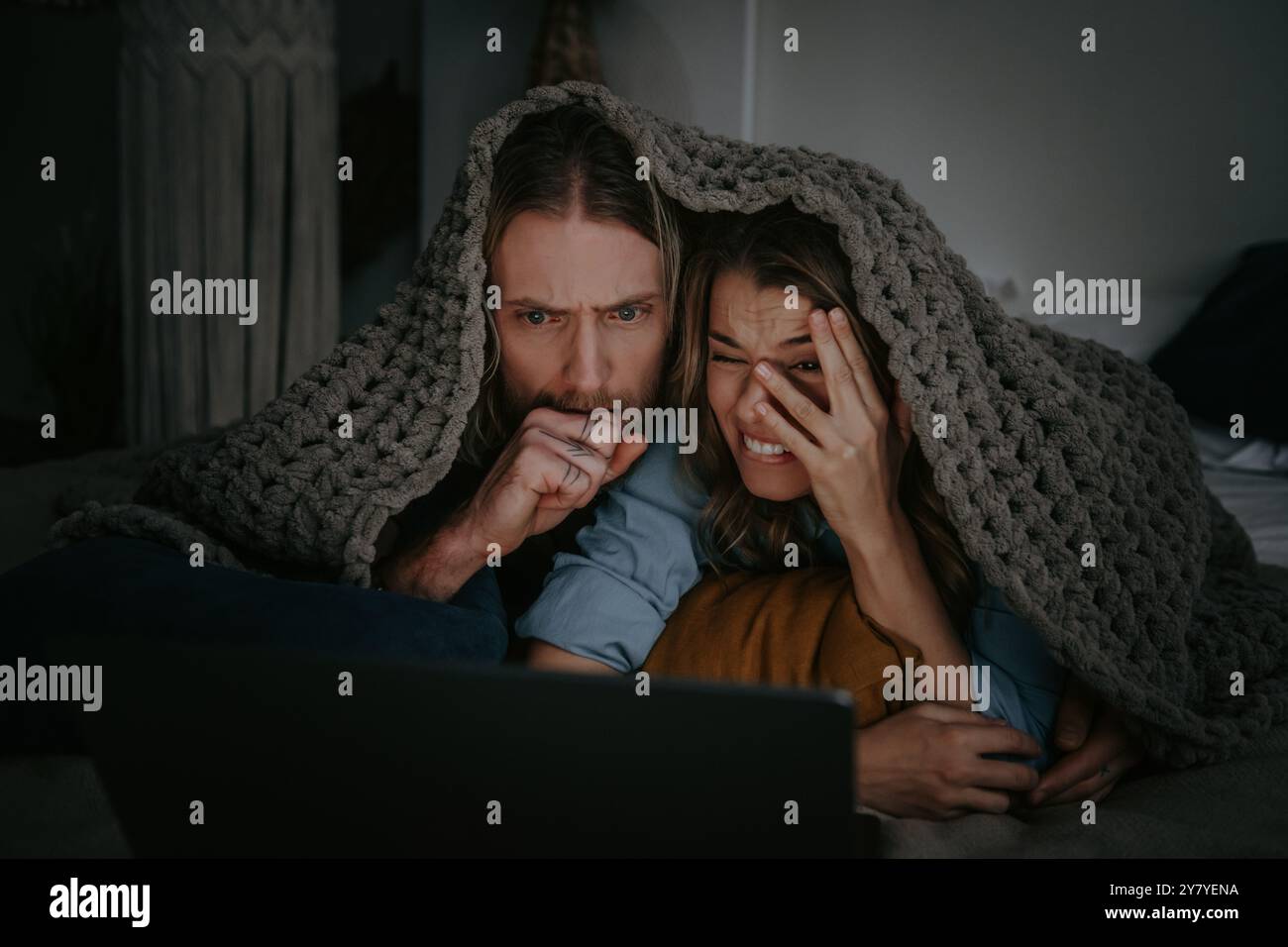 Una giovane coppia spaventata che si copre con una coperta calda e guarda il computer portatile mentre si sdraia a letto a casa insieme Foto Stock Una giovane coppia spaventata che si copre con una coperta calda e guarda il computer portatile mentre si sdraia a letto a casa insieme Foto Stock