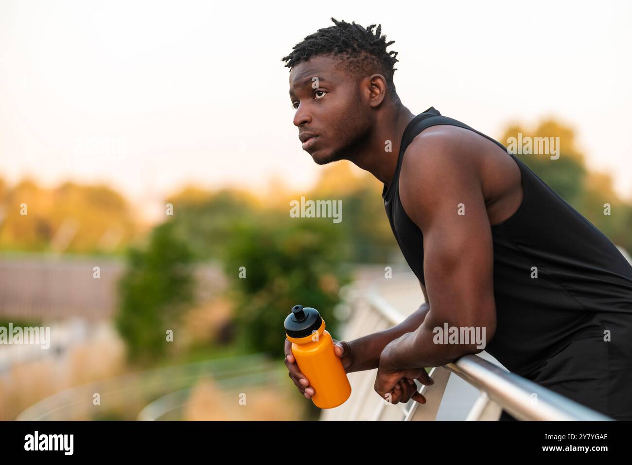 Stanco uomo africano in abbigliamento sportivo che tiene in mano una bottiglia d'acqua mentre riposa dopo un allenamento sportivo all'aperto Foto Stock
