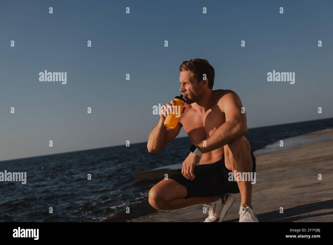 Bell'atleta maschile bevendo acqua e godendosi la vista sul mare dopo gli allenamenti mattutini all'aperto Foto Stock