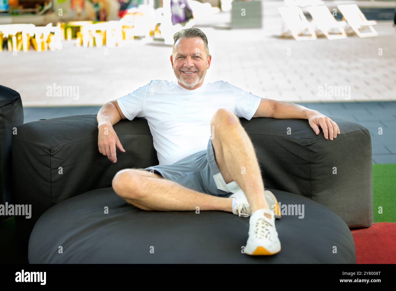 Un uomo di mezza età si sdraia comodamente su un grande divano esterno, vestito con una t-shirt casual bianca, pantaloncini grigi e sneaker. Foto Stock