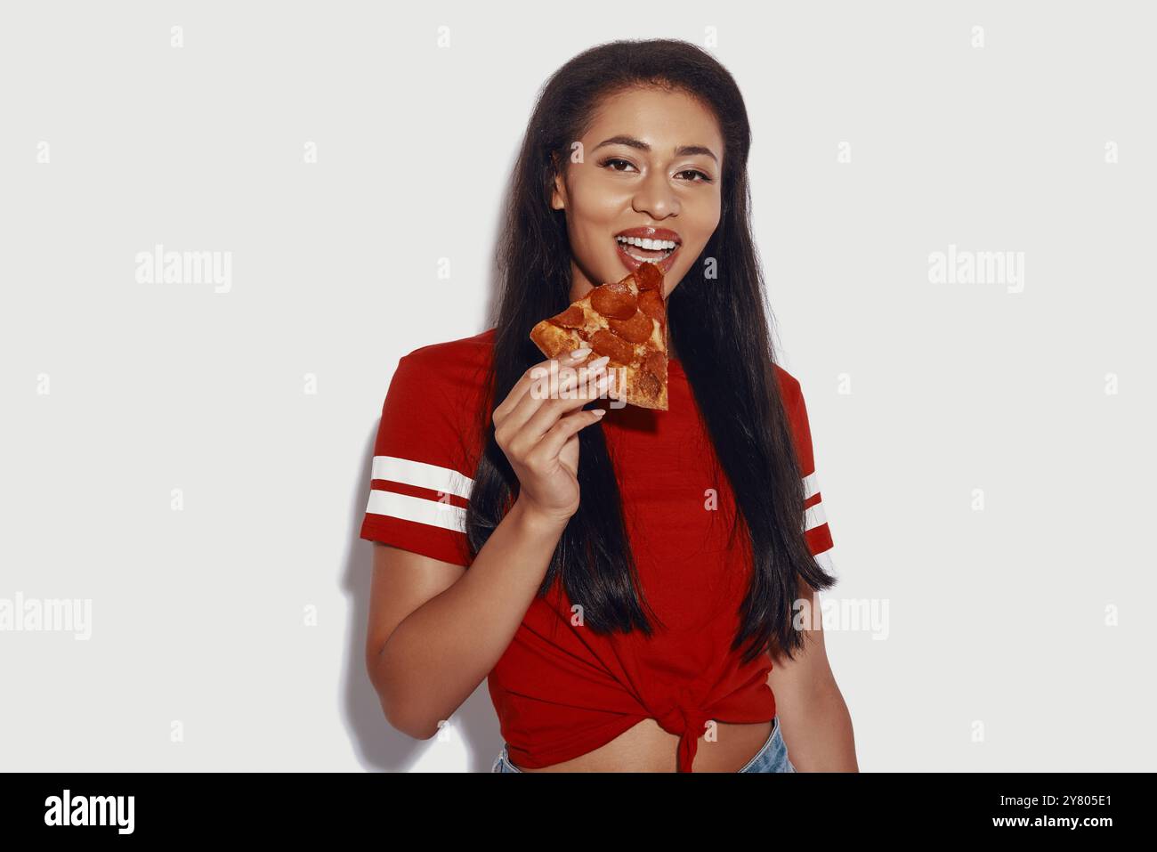 Giovane donna attraente che mangia pizza e guarda la macchina fotografica mentre si trova su uno sfondo grigio Foto Stock