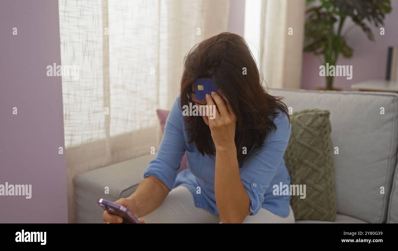 Donna di mezza età turbata che tiene in mano una carta di credito e uno smartphone in salotto, suggerendo problemi finanziari a casa. Foto Stock