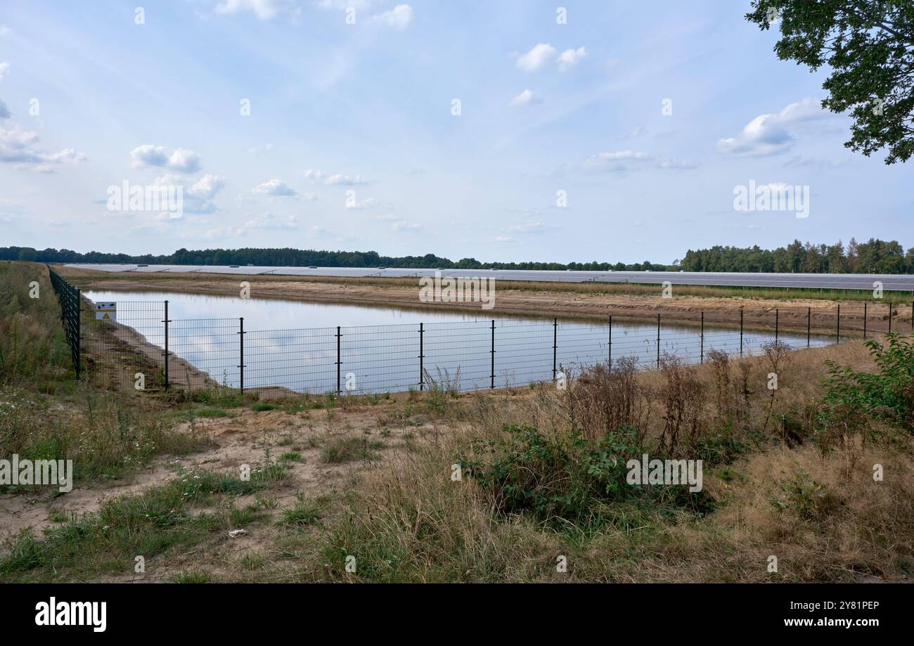 una grande fattoria a energia solare con laghetto circondato da una recinzione con file di pannelli fotovoltaici in un'area rurale con alberi e cieli blu. Rinnovabile e Foto Stock