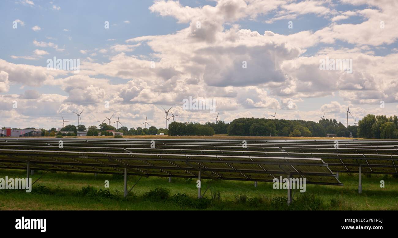 una grande fattoria a energia solare con file di pannelli fotovoltaici, accanto a un'autostrada, ai margini di una città, con alberi, turbine eoliche e cieli nuvolosi. Ren Foto Stock