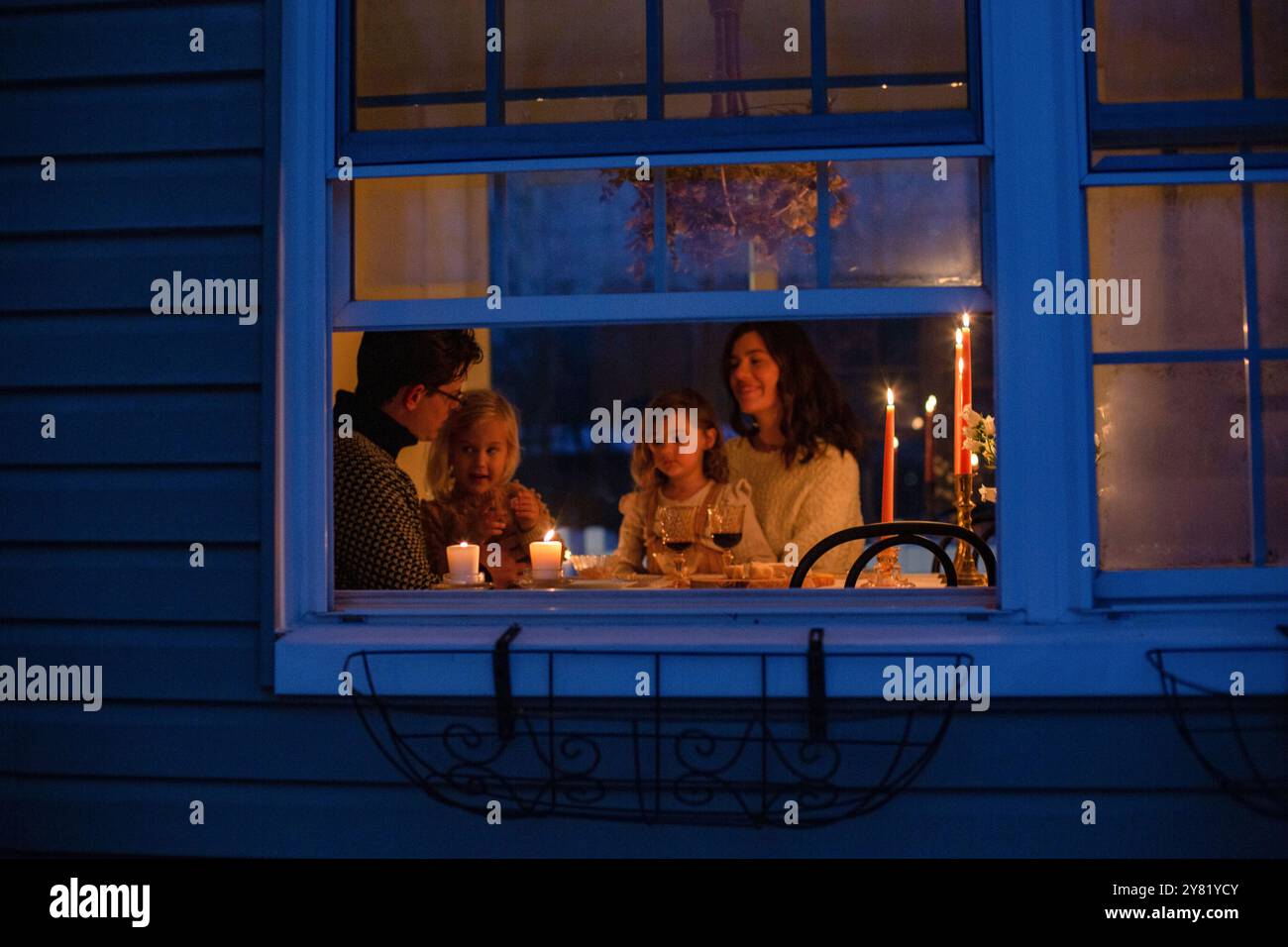 La famiglia può godersi una cena a lume di candela all'interno di una casa accogliente vista dall'esterno della finestra blu illuminata al crepuscolo. Foto Stock