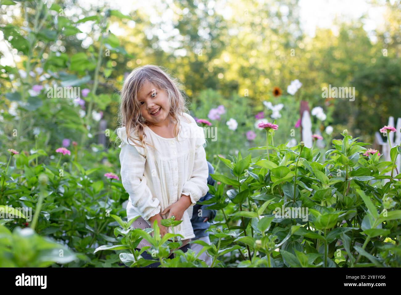 Sorridente ragazza in piedi tra lussureggianti piante verdi in un giardino con fiori che fioriscono sullo sfondo. Foto Stock