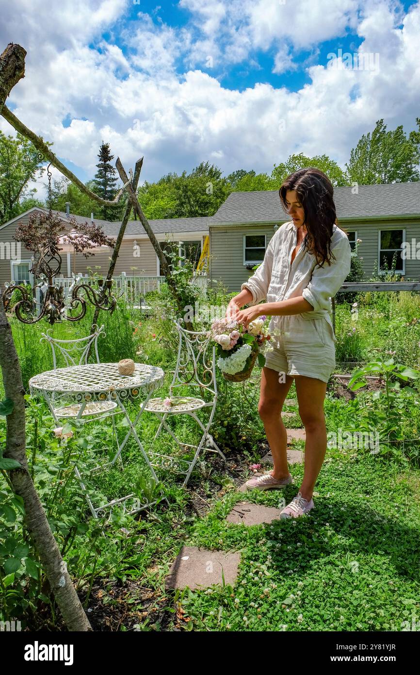 Una donna con un abito estivo tende a fiori in un lussureggiante giardino sul retro con mobili vintage. Foto Stock