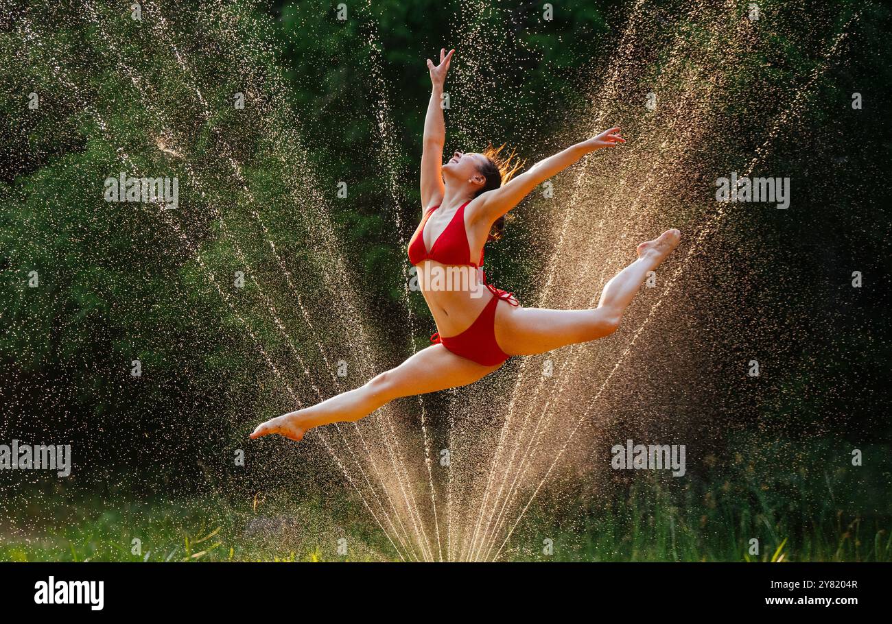 Una graziosa ballerina in un lembo rosso esegue un salto a mezz'aria con spruzzatori d'acqua che creano uno sfondo scintillante. Foto Stock
