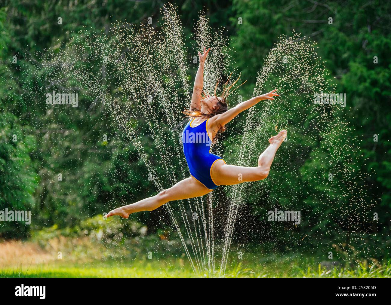 Graziosa ballerina che salta sopra gli irrigatori d'acqua in un lussureggiante parco verde. Foto Stock