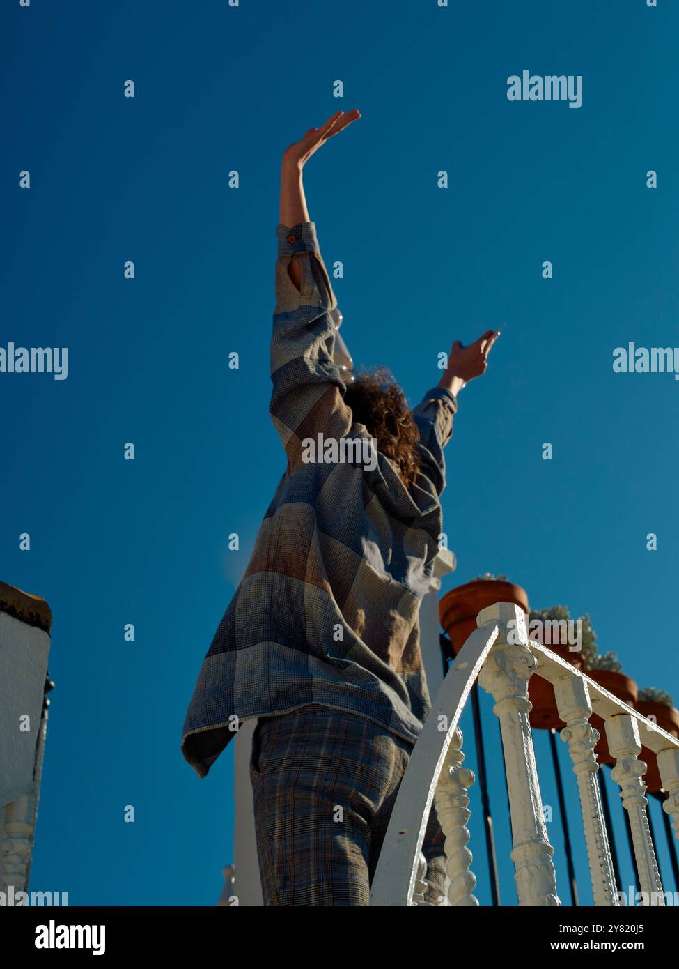 Una persona estende le braccia verso il cielo in piedi da una ringhiera bianca su uno sfondo blu chiaro. Foto Stock