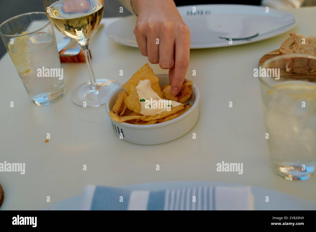 La mano di una persona prende una scheggia con un tuffo da una ciotola su un tavolo da pranzo con un bicchiere di vino bianco e pane sullo sfondo. Foto Stock