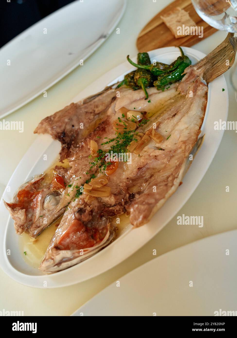 Un pesce appena cotto su un piatto ovale bianco guarnito con erbe e accompagnato da peperoni verdi. Foto Stock