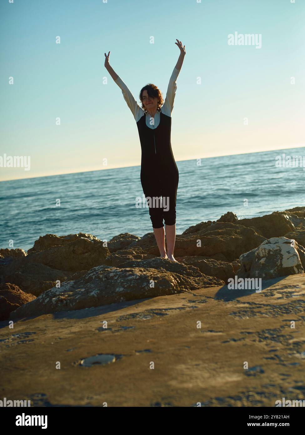 Donna con le braccia alzate in piedi sulla costa rocciosa sullo sfondo dell'oceano al tramonto Foto Stock