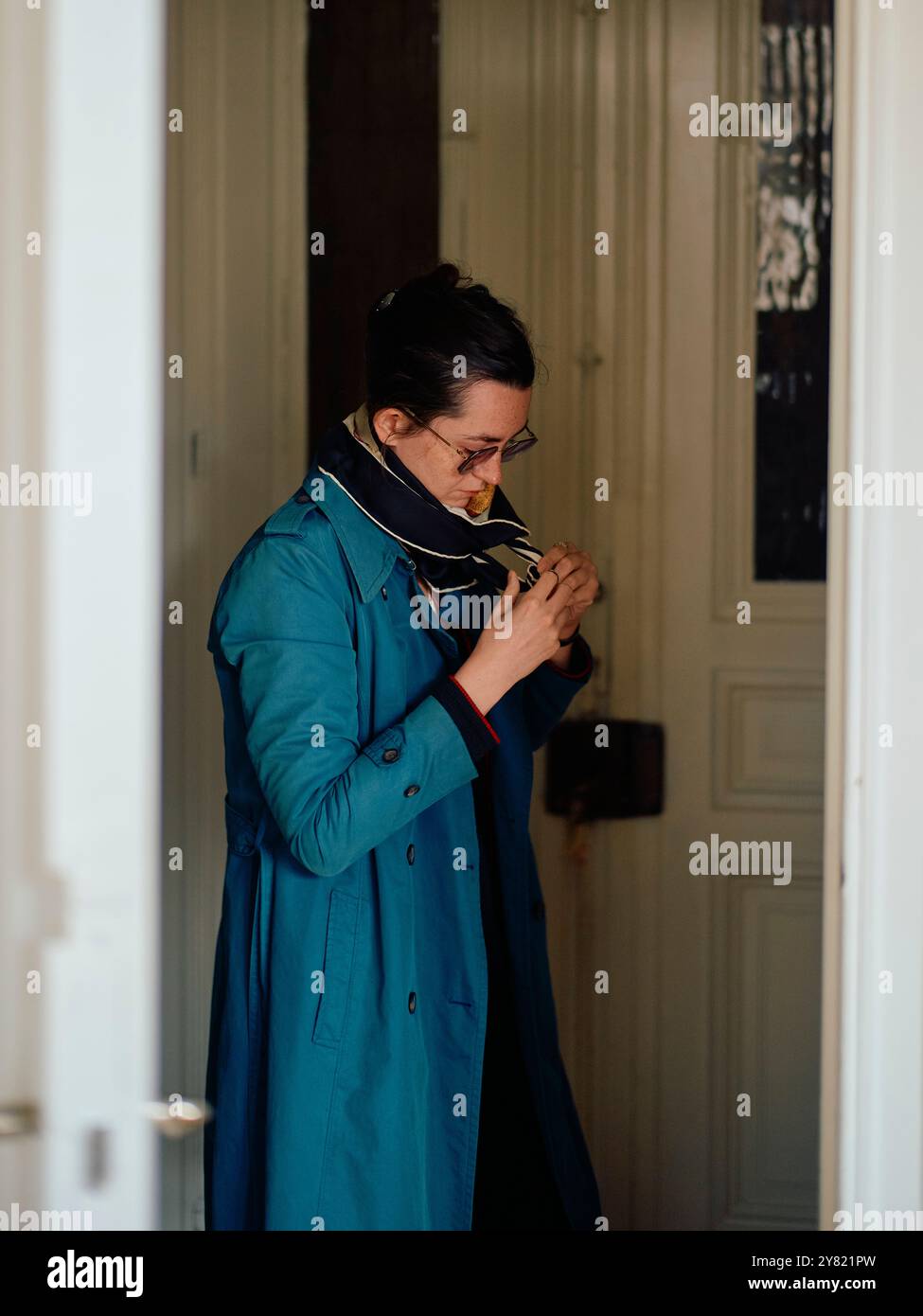 Donna con un cappotto di tè che accende una sigaretta all'interno di una porta con una porta bianca Foto Stock