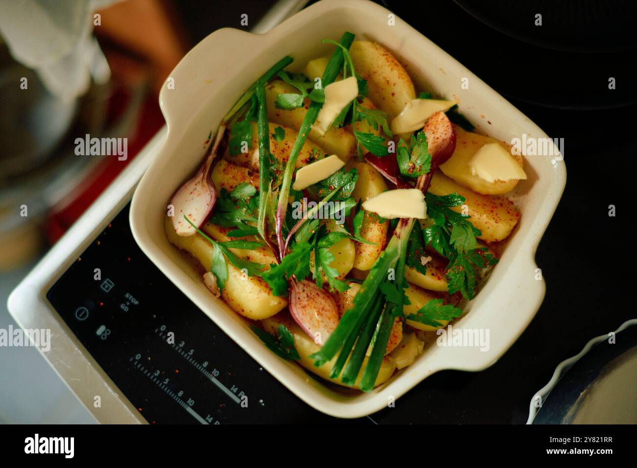 Spicchi di patate al forno guarniti con prezzemolo fresco e scaglie di parmigiano in un piatto bianco da forno su una stufa a induzione. Foto Stock