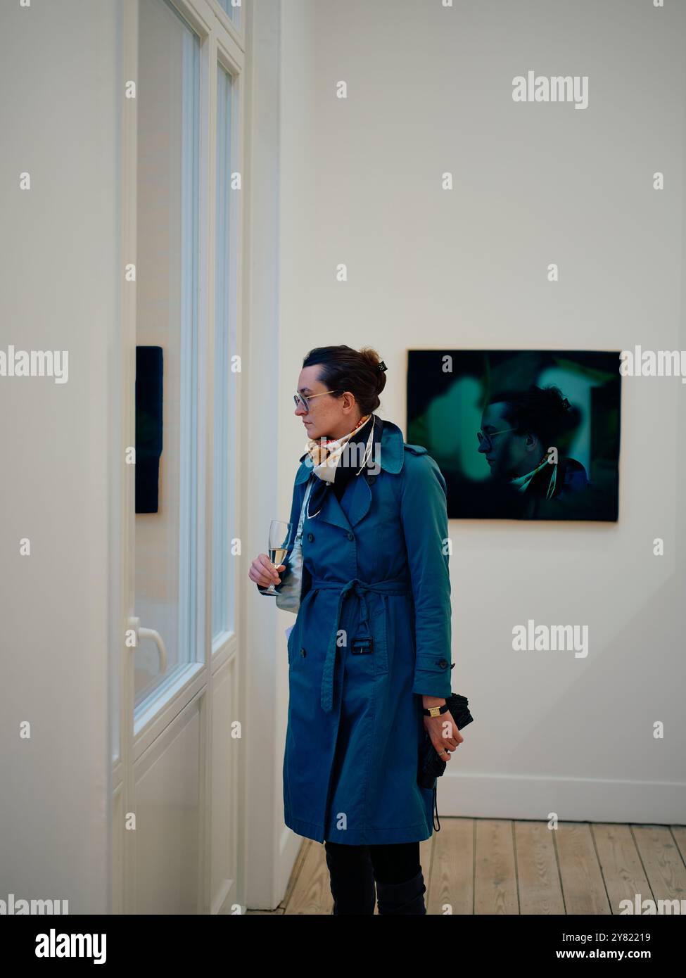 Una donna con un cappotto color verde acqua con le cuffie si trova in una galleria d'arte a guardare una fotografia. Foto Stock