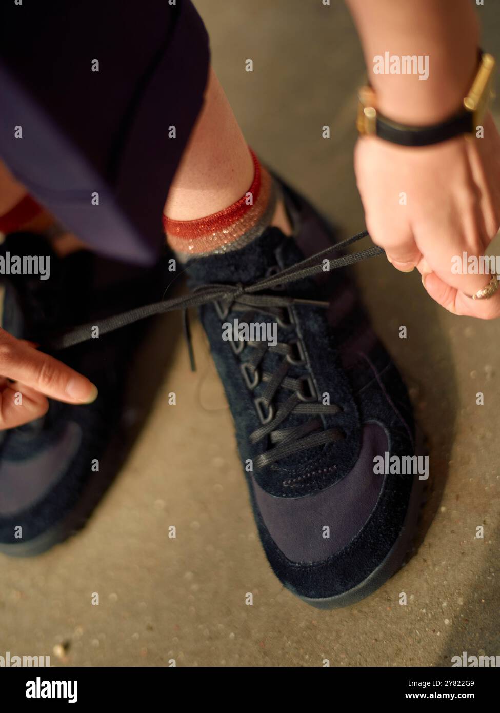 Una persona sta legando i lacci delle sue scarpe sportive blu navy con le mani adornate da bracciali dorati e da un luccicante orologio nero. Foto Stock