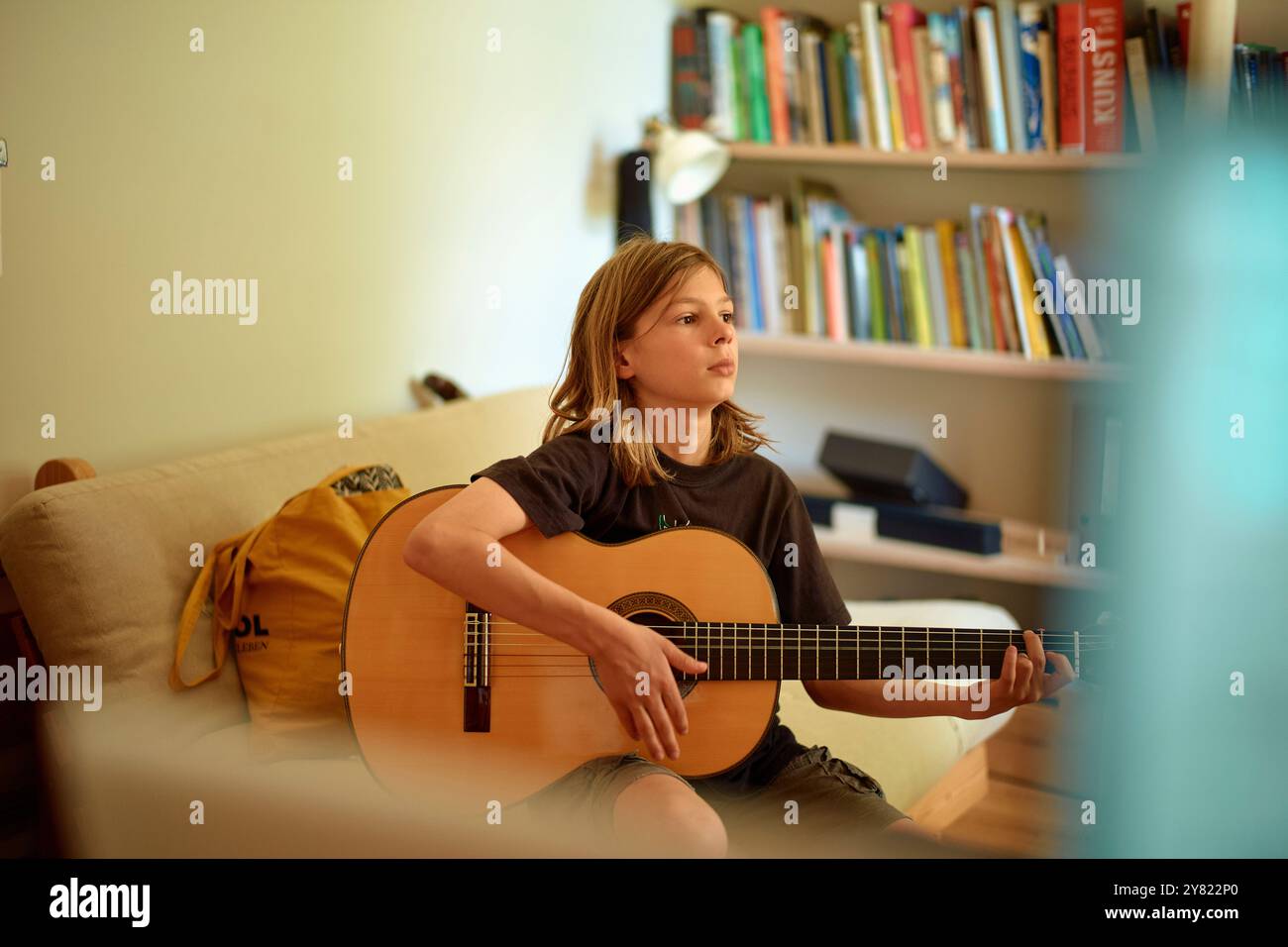 Giovane musicista che suona una chitarra acustica in una stanza con una libreria sullo sfondo. Foto Stock
