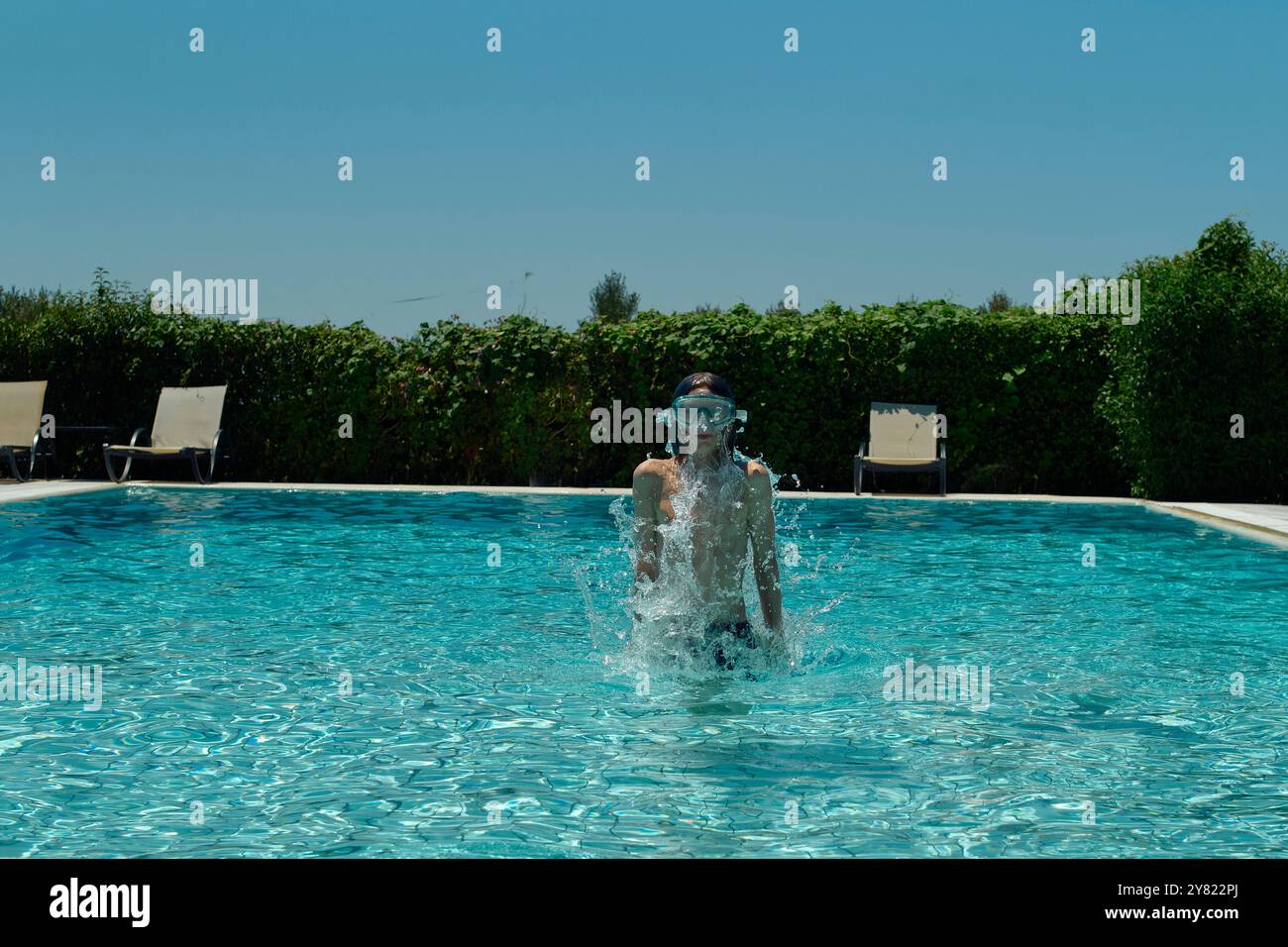 Una persona con gli occhiali da bagno esplode energicamente attraverso la superficie di una piscina blu chiaro in una giornata di sole, con sedie a sdraio e siepi verdi sullo sfondo. Foto Stock