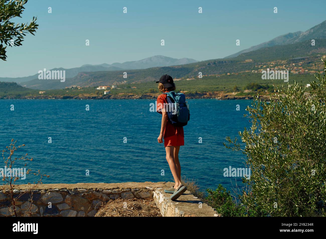 Viaggiatore solo si affaccia su un mare blu tranquillo con montagne in lontananza, adagiato su una sporgenza di pietra mentre indossa uno zaino e un cappello da sole. Foto Stock