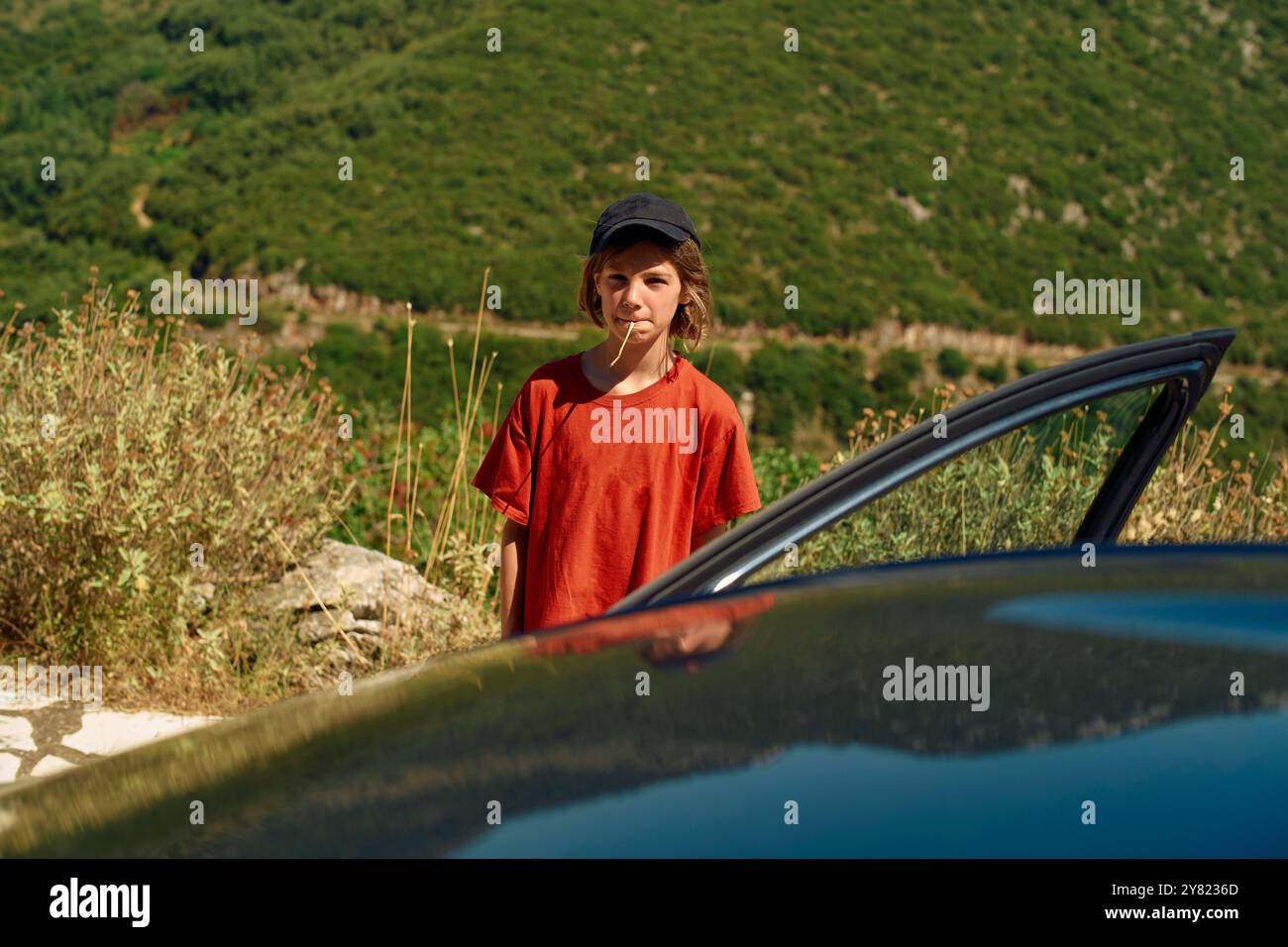 Un bambino con una camicia rossa e un berretto nero si trova accanto alla porta di una macchina con un lecca lecca in bocca, affacciato su una lussureggiante collina verde. Foto Stock