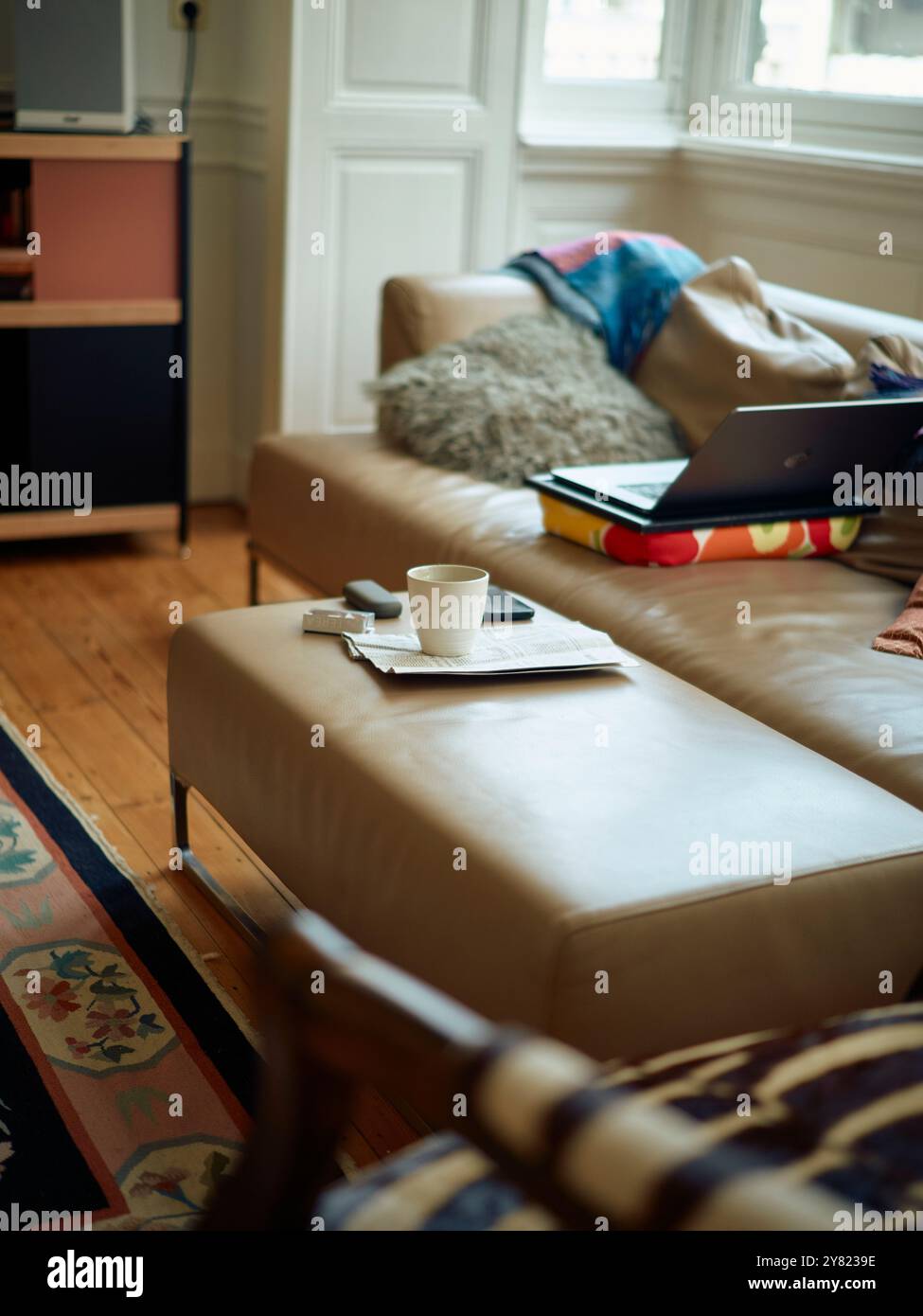 Un comodo sistema di home Office con un computer portatile su una scrivania colorata, una tazza di caffè sull'ottomana e un comodo spazio abitativo sullo sfondo. Foto Stock