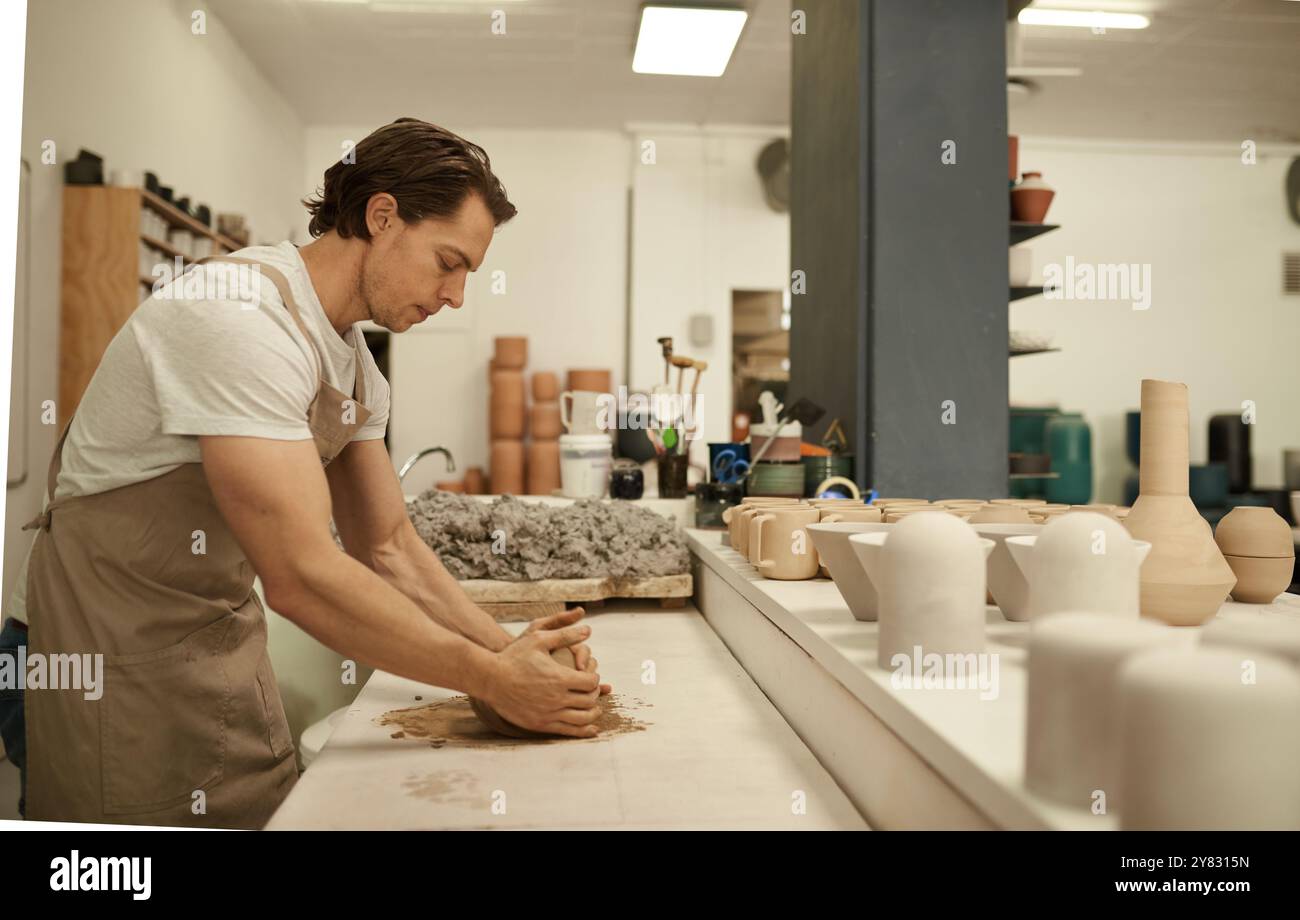 Giovane vasaio maschile che indossa un grembiule che forma un pezzo di argilla bagnato mentre lavora a un tavolo in uno studio di ceramica Foto Stock