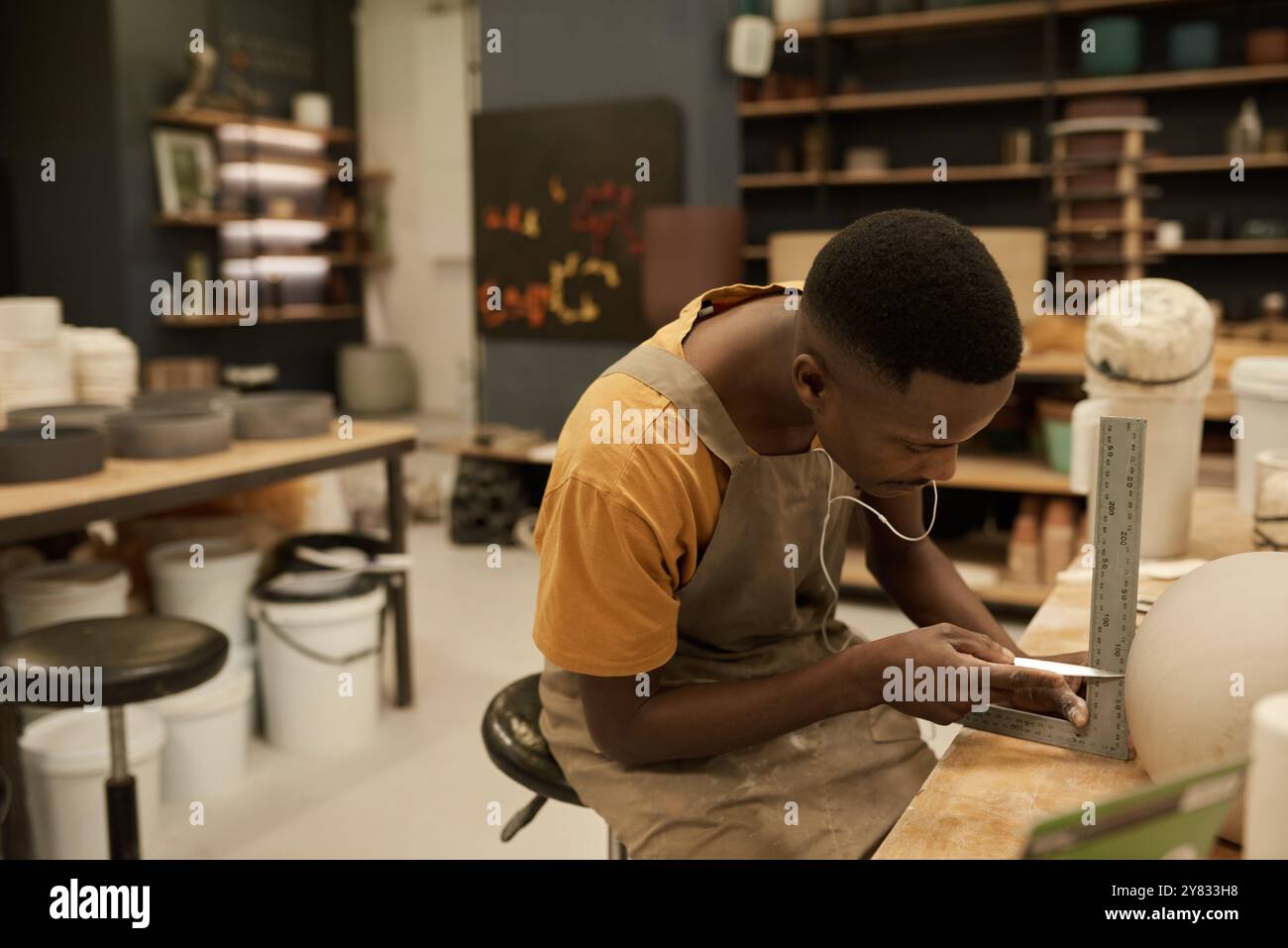 Giovane ceramista africano che usa lo strumento e il righello per segnare un buco in una pentola di argilla su una panchina in uno studio di ceramica Foto Stock