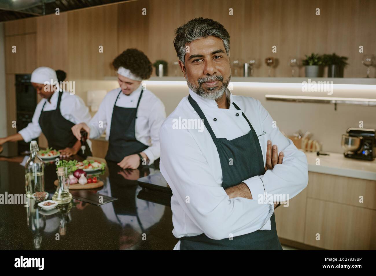 Ritratto dello chef indiano maturo in uniforme bianca sorridendo leggermente mentre posa per una foto nella cucina del proprio ristorante Foto Stock