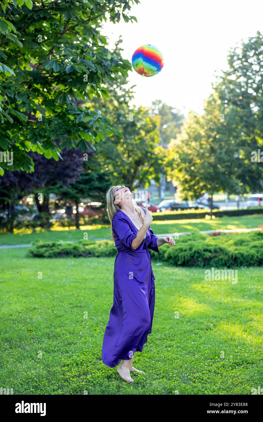 Bella donna con capelli lunghi e abito viola si sta godendo nel gioco nel parco pubblico con palla colorata. Foto Stock