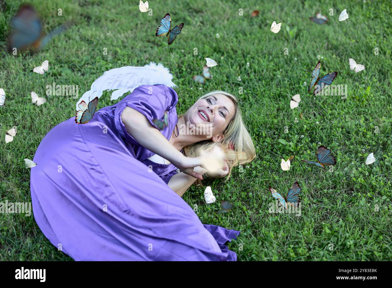 La bella bionda con capelli lunghi e il lungo abito viola ha le ali bianche e sembra un angelo. È sdraiata nel parco pubblico durante una calda S Foto Stock
