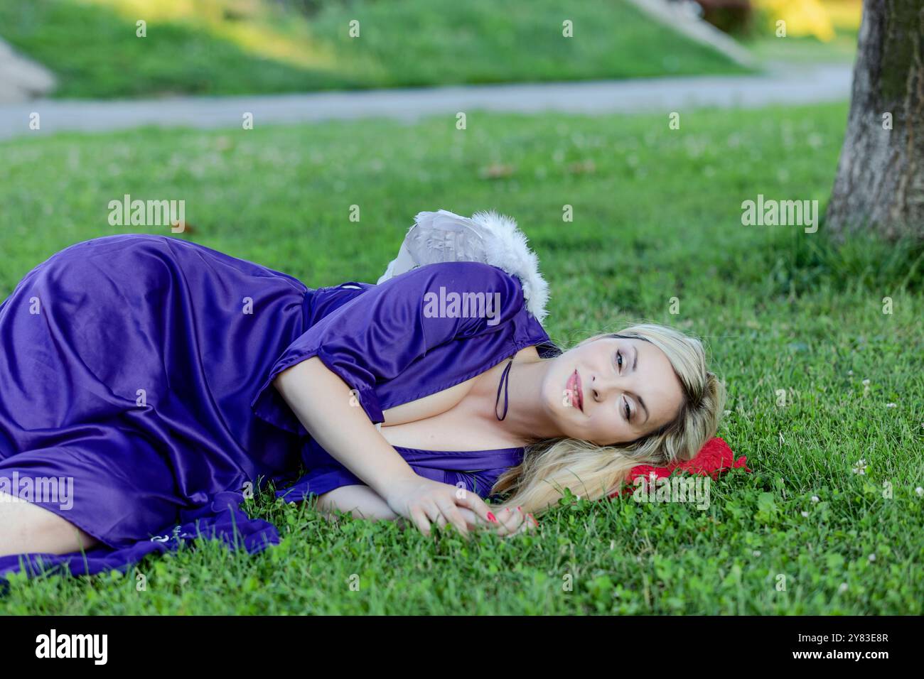La bella bionda con capelli lunghi e il lungo abito viola ha le ali bianche e sembra un angelo. Sta camminando nel parco pubblico durante un caldo Foto Stock