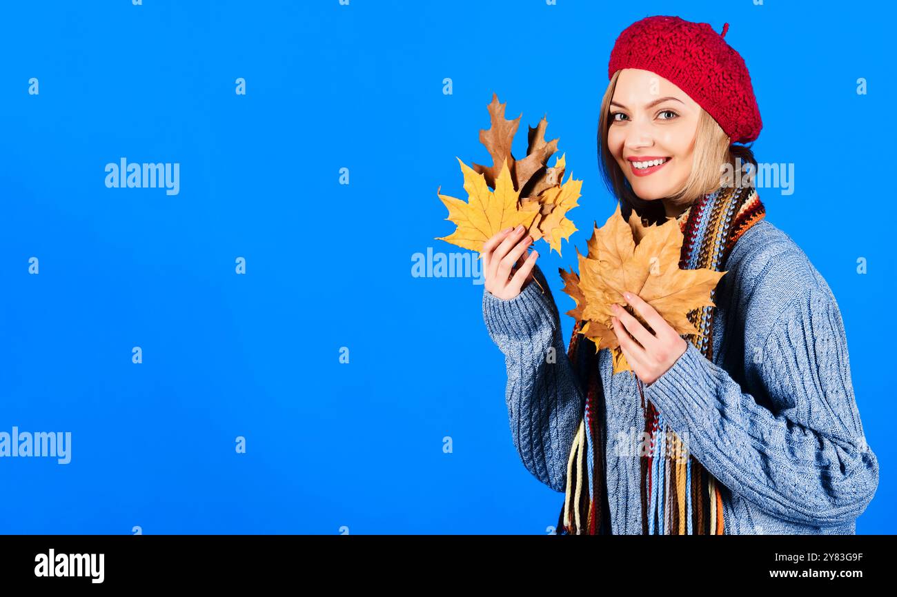 Donna autunnale sorridente in maglione grigio, sciarpa colorata e cappello rosso con foglie autunnali. Elegante ragazza in un abbigliamento alla moda con foglie autunnali. Moda autunnale Foto Stock