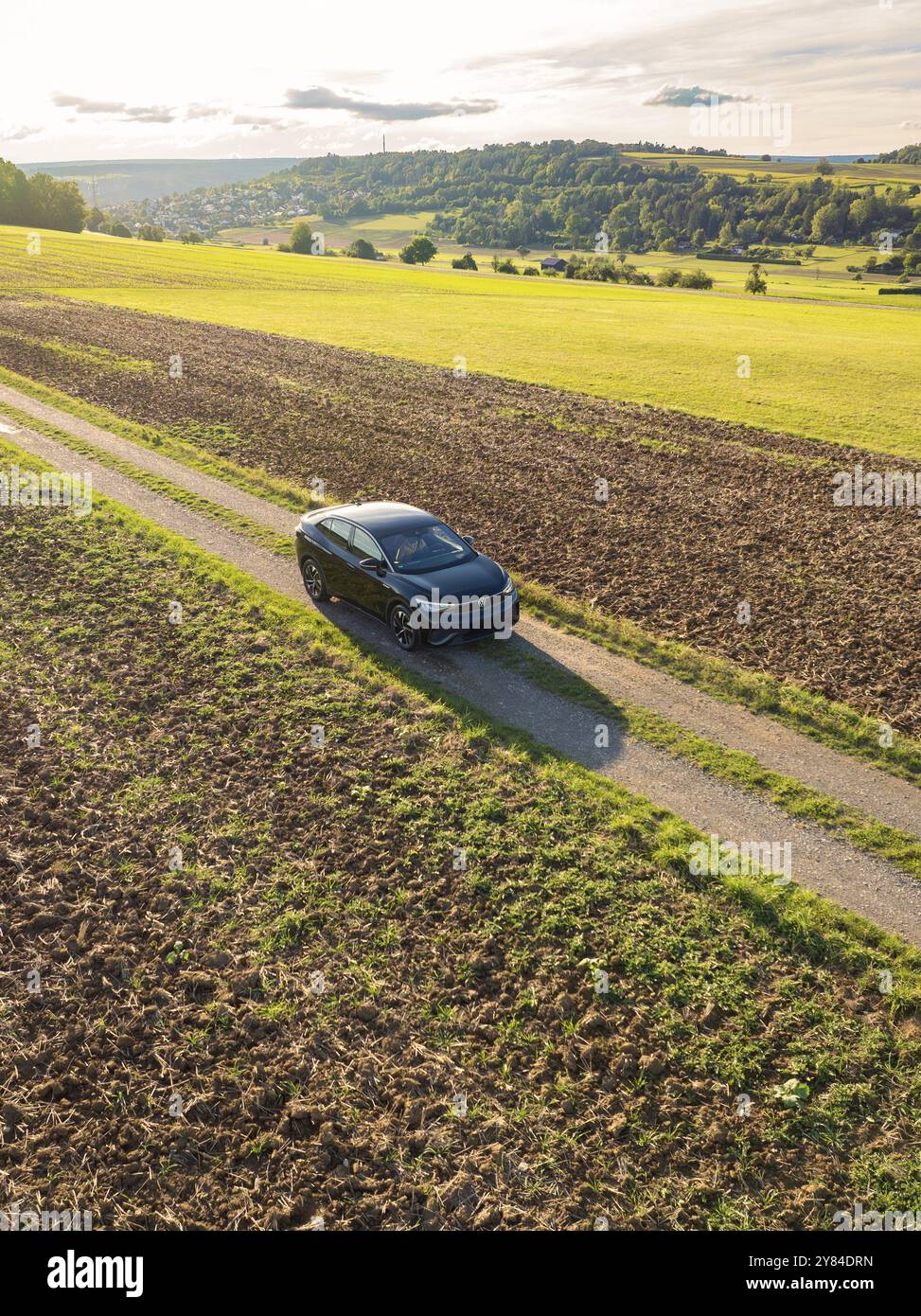 Un'auto guida su una pista sterrata tra grandi campi sotto un cielo con nuvole e sole serale, car sharing, VW ID5, Calw, Foresta Nera, Germania, Europa Foto Stock