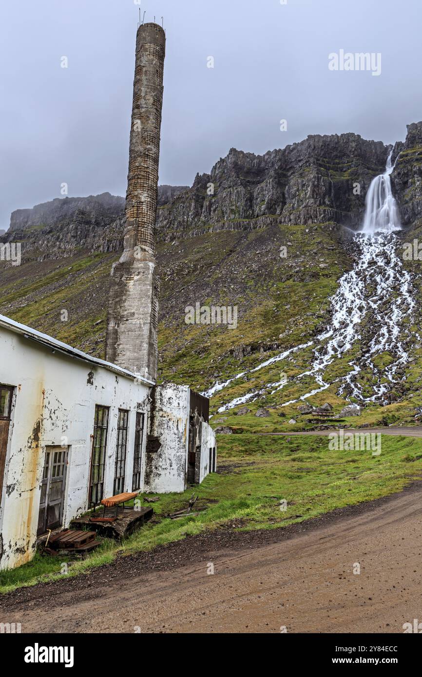 Vecchia fabbrica, fabbrica di pesce, camino, in disuso, cascata e scogliere dietro, nuvoloso, Djupavik, Strandir, Westfjords, Islanda, Europa Foto Stock