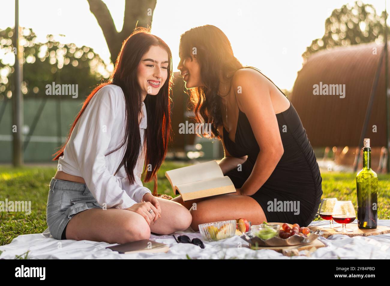 due amici latini sorridenti e felici mentre si godono il picnic, parlano e raccontano segreti nelle orecchie dell'altro, tramonta sullo sfondo. Foto Stock