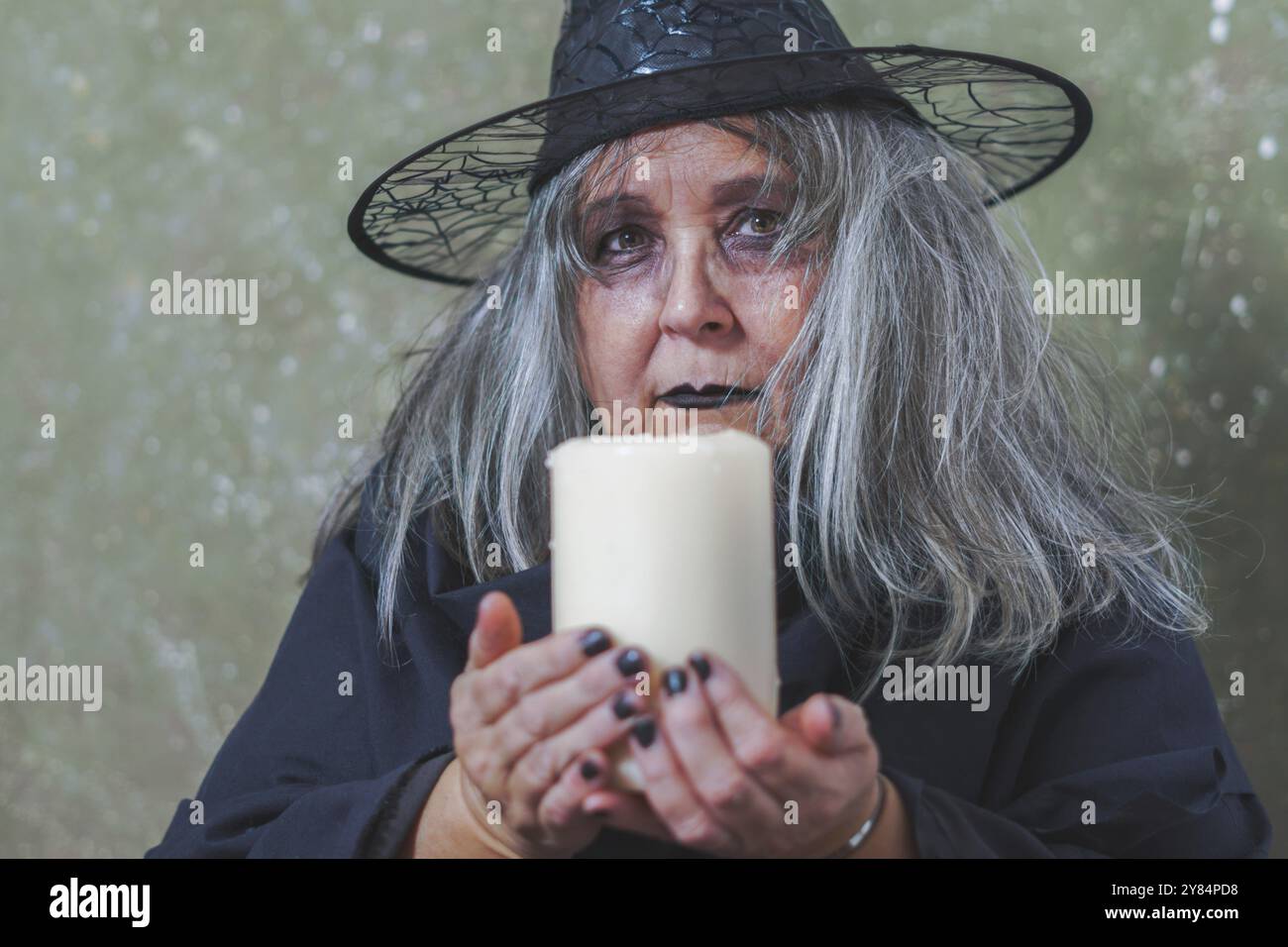 Vecchia strega dai capelli bianchi vestita di nero con un cappello e una grande candela bianca nelle sue mani Foto Stock