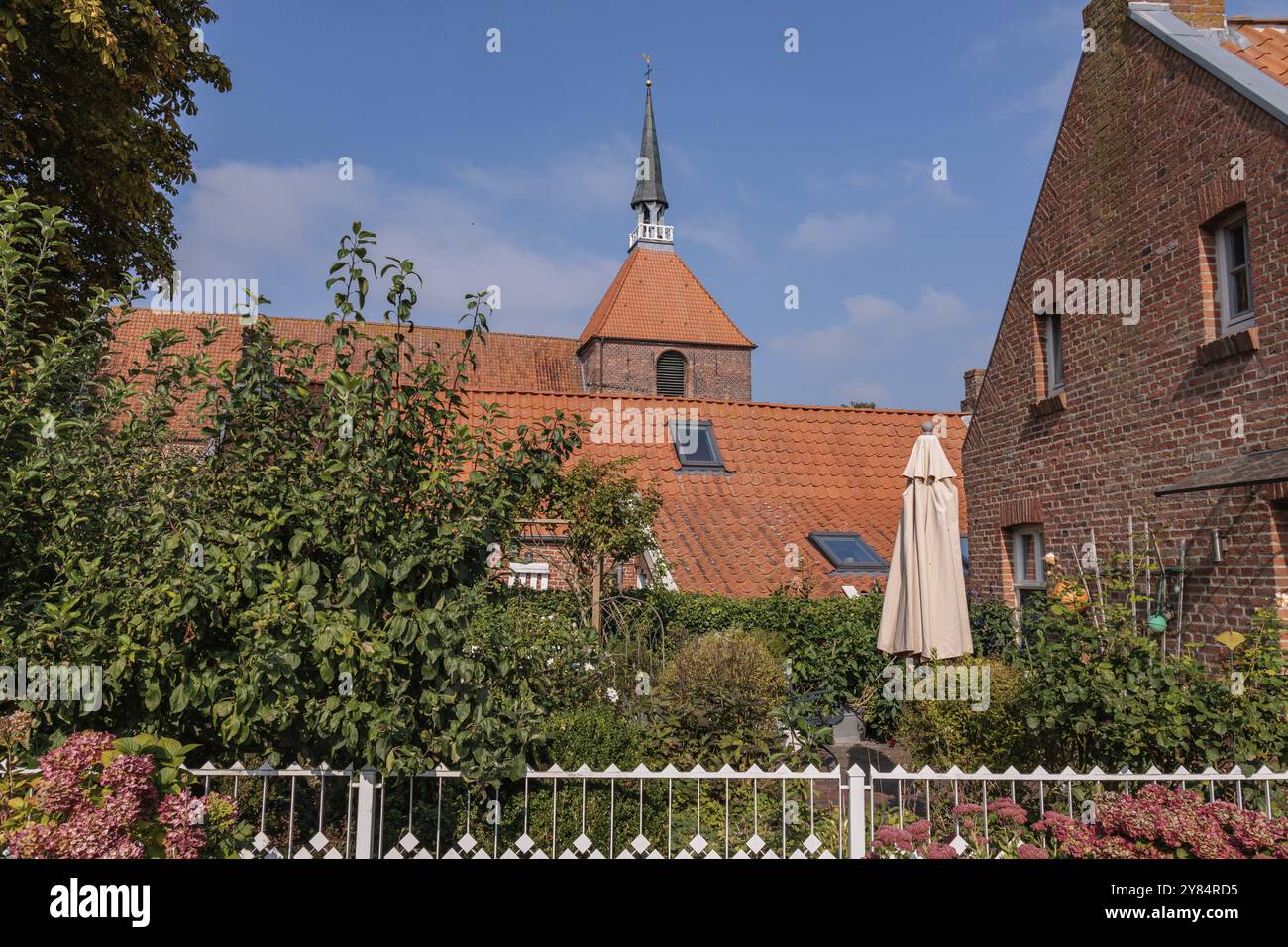 Un bellissimo giardino con la cima di una chiesa in mattoni e un cielo soleggiato sullo sfondo, krummhoern, frisia orientale, germania Foto Stock