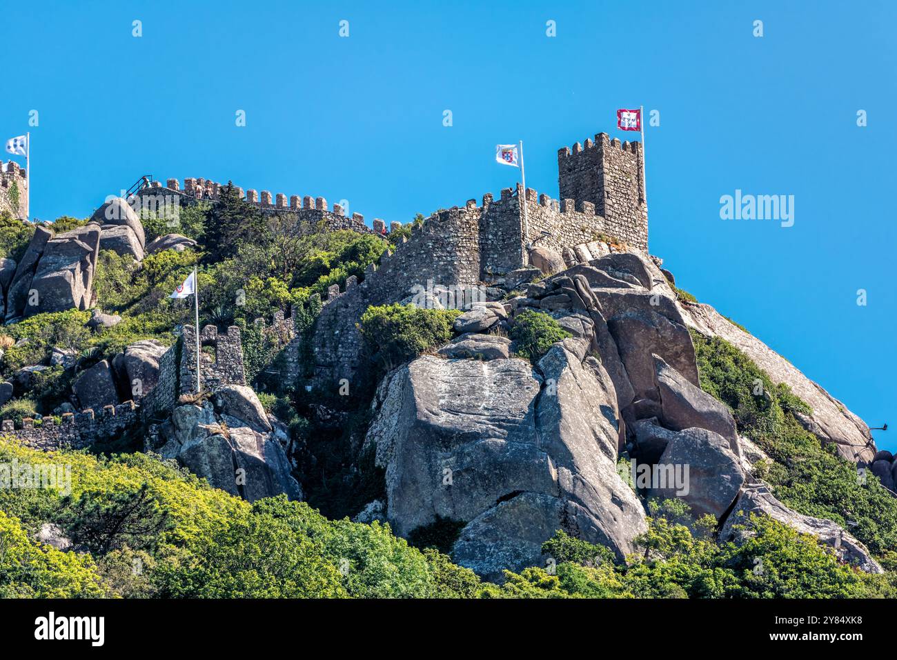 Castello Moresco Castelo Dos Mouros Sintra Portogallo | SINTRA, Portogallo — I visitatori percorrono il camminamento restaurato del Castelo dos Mouros mentre sale lungo la cresta di granito sopra Sintra, le bandiere della fortezza segnano le sue torri contro un cielo sereno. I sovrani islamici costruirono la fortezza tra il IX e l'XI secolo per difendere gli accessi a Lisbona, mantenendola fino al 1147, quando Afonso Henriques conquistò Sintra dopo la conquista di Lisbona. Foto Stock
