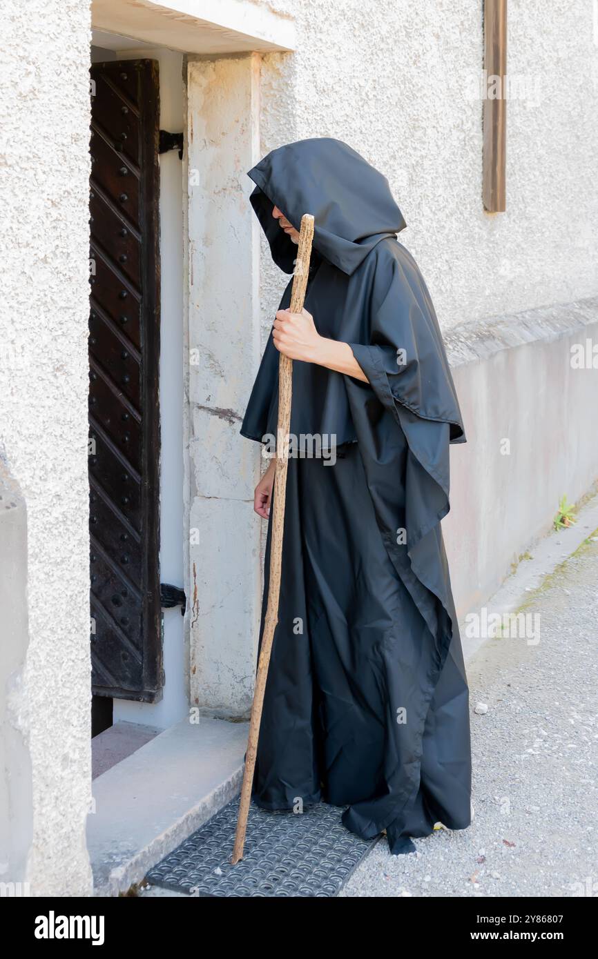Un monaco anziano con un bastone in una cazzuola nera cammina attraverso le porte aperte di una vecchia chiesa. Foto Stock