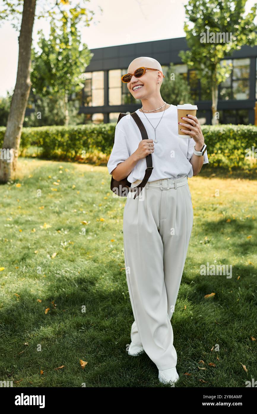 Una donna calva sorride brillantemente mentre passeggia attraverso un parco vivace con un drink in mano. Foto Stock