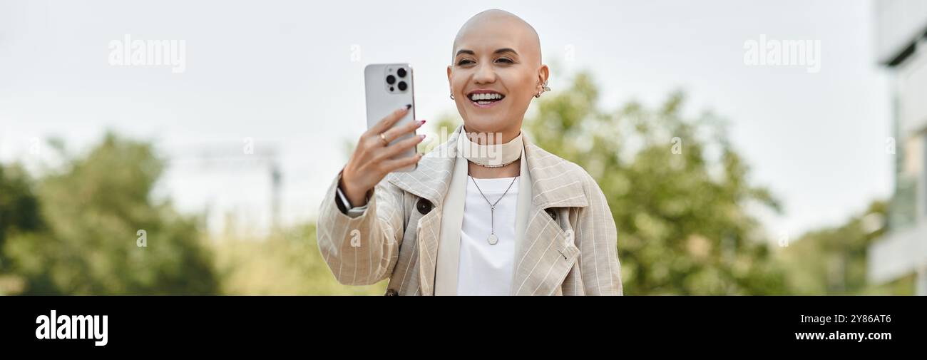 Una giovane donna calva vestita elegantemente cattura un gioioso selfie all'aperto alla luce del sole. Foto Stock