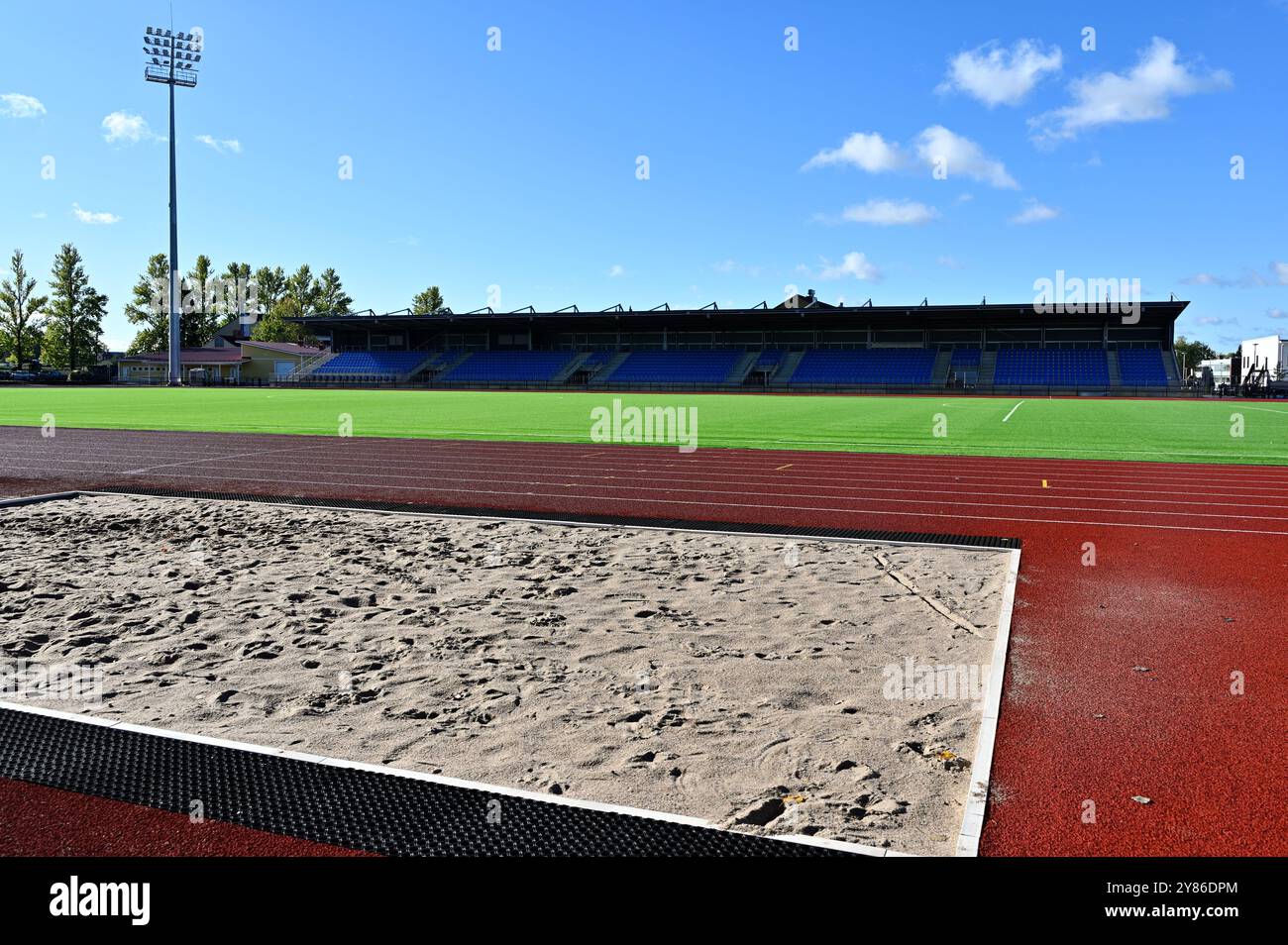 Questa immagine raffigura uno stadio di atletica sotto un cielo azzurro, che mostra un lungo salto di sabbia in primo piano, una pista da corsa rossa e un vivace gr Foto Stock