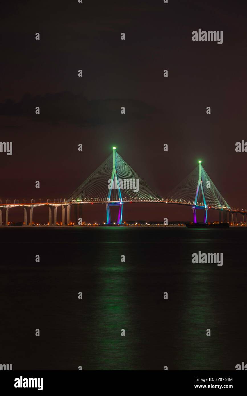 Una vista notturna di un ponte splendidamente illuminato, caratterizzato da luci blu e viola che si riflettono sull'acqua calma, creando un'atmosfera serena. Foto Stock