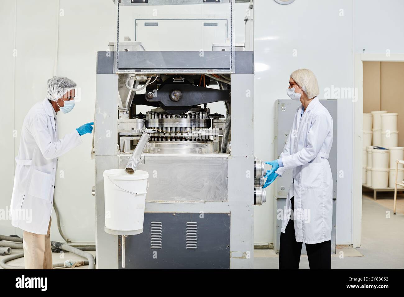 Vista laterale di due tecnici di processo in camici e maschere da laboratorio che ispezionano la pressa per tablet e monitorano la produzione di farmaci nello stabilimento di produzione farmaceutica Foto Stock