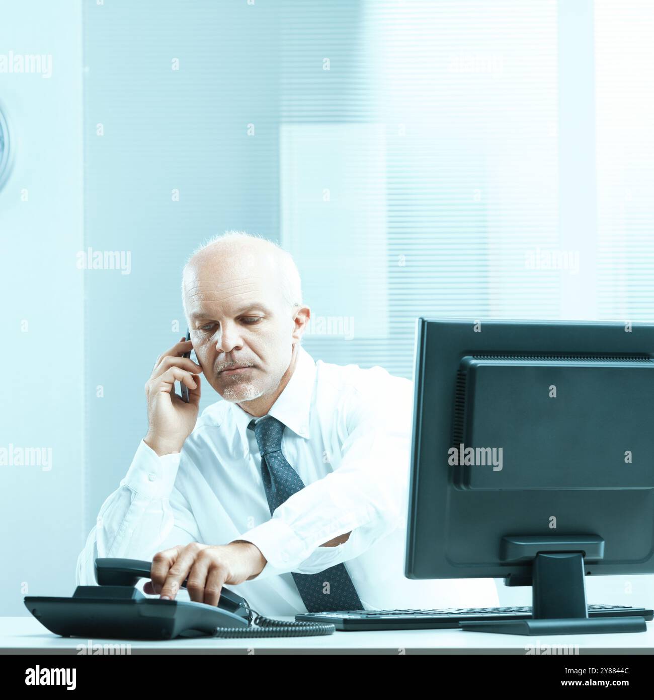 Un uomo d'affari maturo è seduto alla sua scrivania in ufficio, a fare una telefonata e a comporre un numero su un telefono fisso. Indossa una camicia bianca e.. Foto Stock