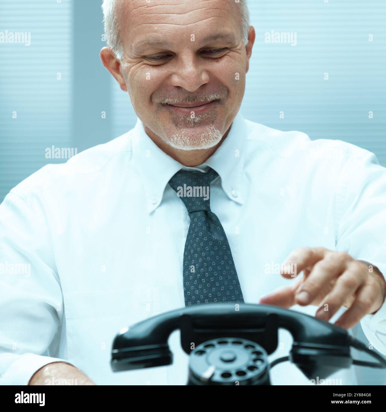 Un uomo d'affari maturo con una camicia bianca sorride mentre compone un telefono vintage alla sua scrivania, mescolando tecnologia vecchia e nuova Foto Stock