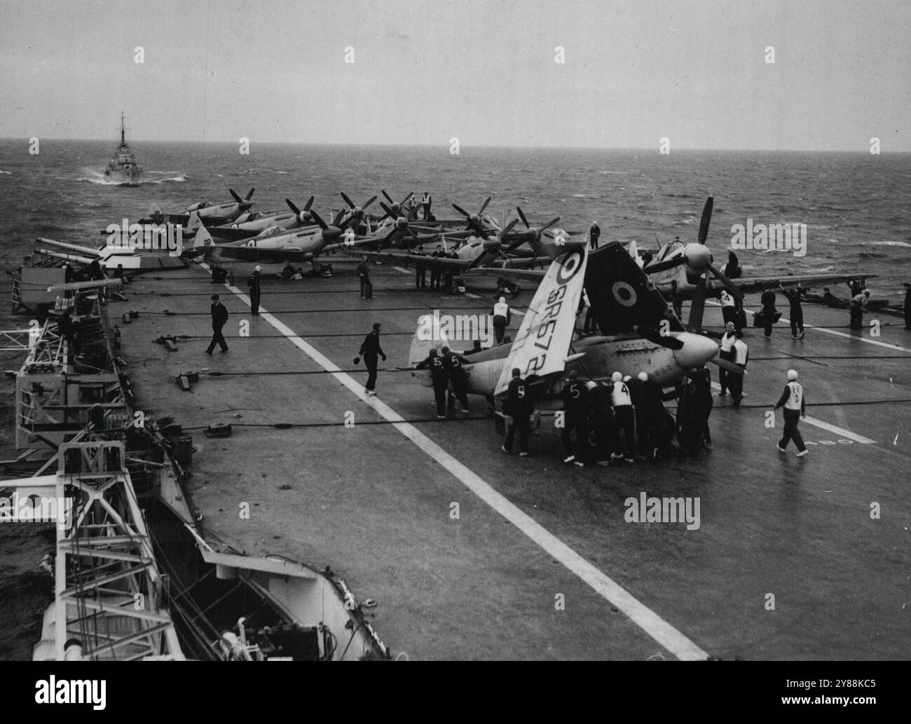 Addestramento "Illustrious" per i velivoli della marina pert-time -- Un aereo da caccia Seafire di uno degli squadroni viene spinto in posizione sul ponte di volo dell'"Illustrious" prima dei voli di addestramento. Sullo sfondo si può vedere il cacciatorpediniere di stanza, H.M.S. Roebuck. Gli squadroni Seafire di tre squadroni aerei della Royal Naval Volunteer Reserve sono in fase di addestramento a bordo della portaerei "Illustrious" durante le crociere nel Canale al largo della costa sud-occidentale della Manica. In tutto 26 aerei furono imbarcati per l'addestramento di quindici giorni, e gli uomini che vi parteciparono includono ufficiali non volanti e manutentori aerei Foto Stock